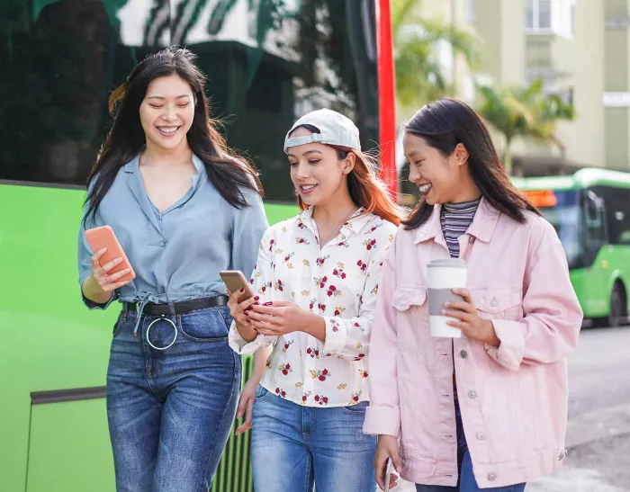 3 girls looking at smartphones and laughing in front of a bus