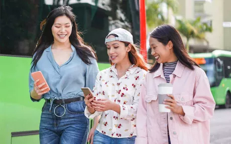 3 girls looking at smartphones and laughing in front of a bus